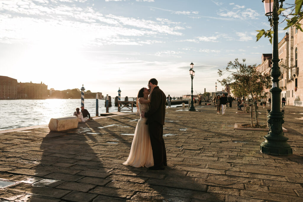 Couple embracing on the sunlit Zattere waterfront in Dorsoduro, Venice, with long shadows across the stone pavement and the Giudecca Canal beside them.