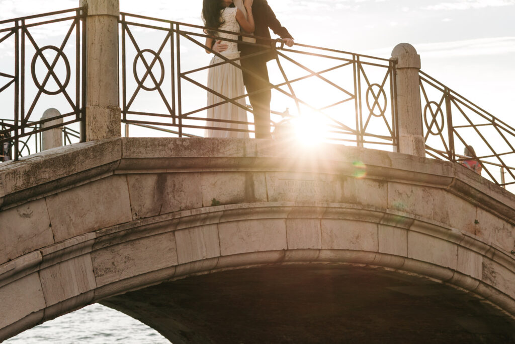 Couple standing close together on an arched stone bridge in Venice, with sunlight streaming through the decorative iron railing.