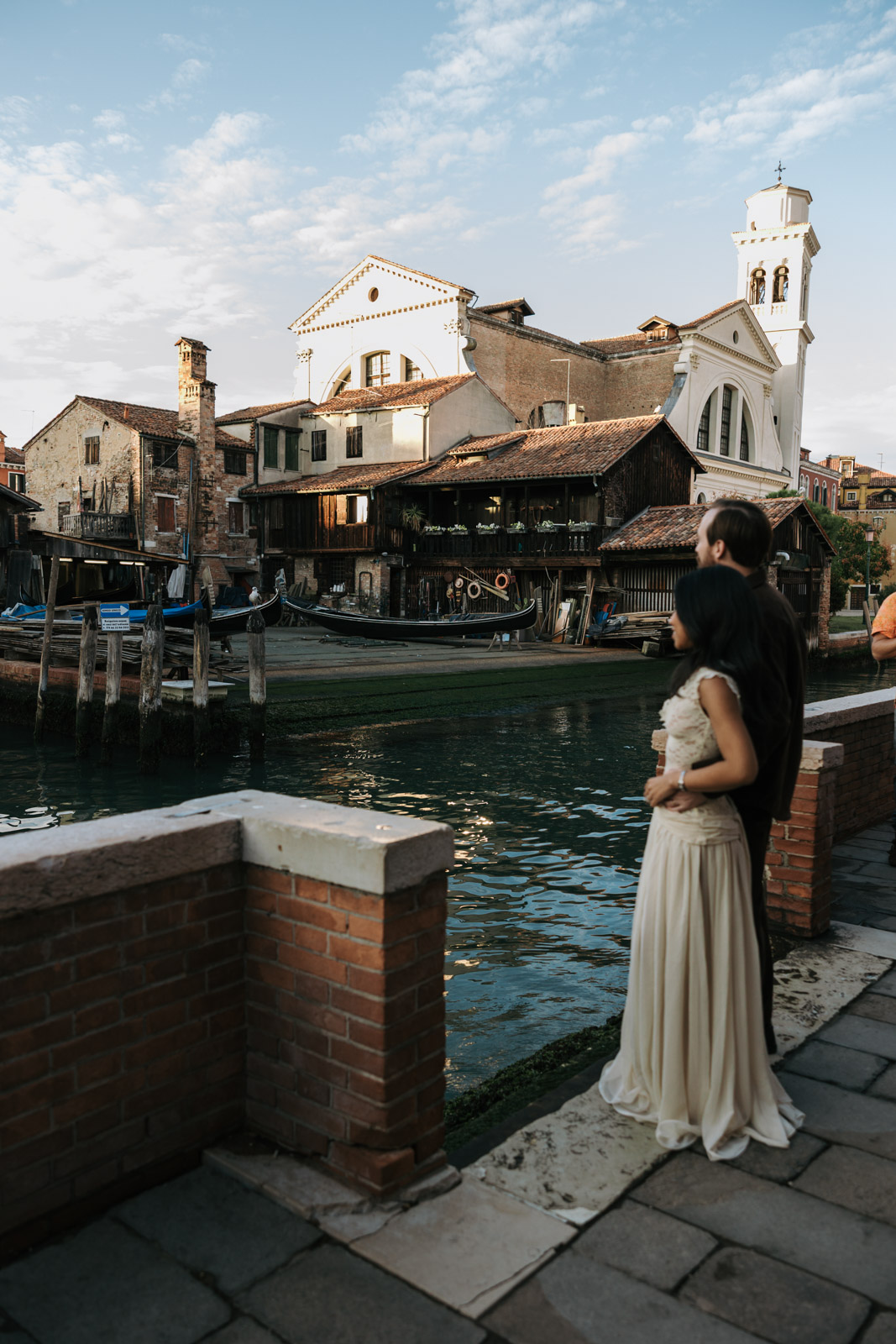 Couple embracing beside a canal opposite Squero di San Trovaso in Venice, with gondolas and wooden boatyard buildings in the background.