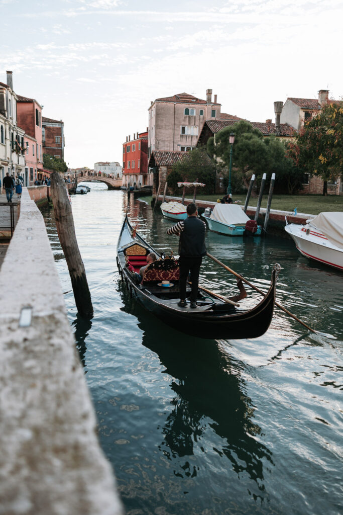 Gondolier rowing a black gondola along a quiet canal in Dorsoduro, Venice, past moored boats and colourful waterfront buildings.