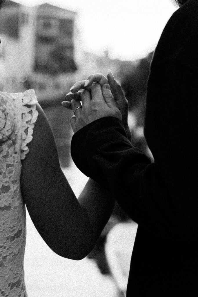 Black-and-white close-up of a couple holding hands, with her floral lace dress beside his dark sleeve.