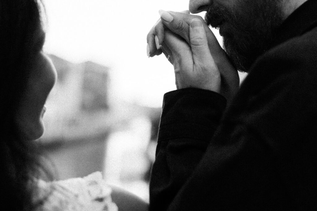 Black-and-white close-up of a man gently kissing his partner’s hand, with her lace dress and blurred Venetian buildings in the background.