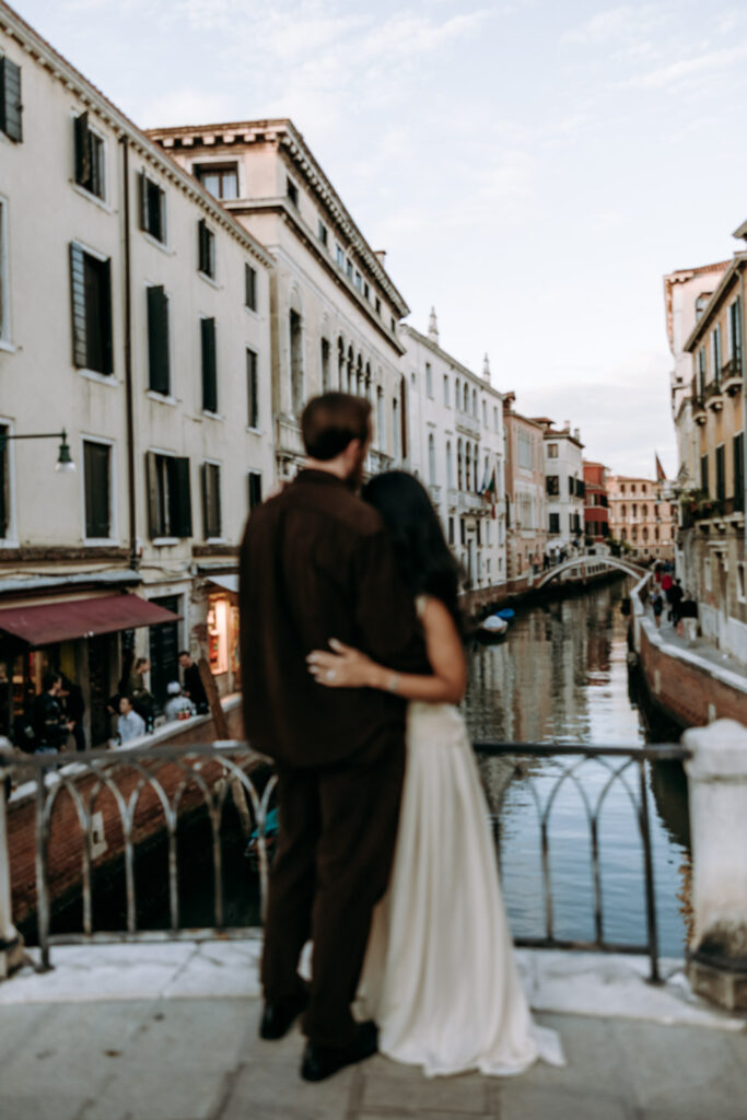 Couple seen from behind embracing on a bridge in Dorsoduro, Venice, overlooking a narrow canal lined with historic buildings.