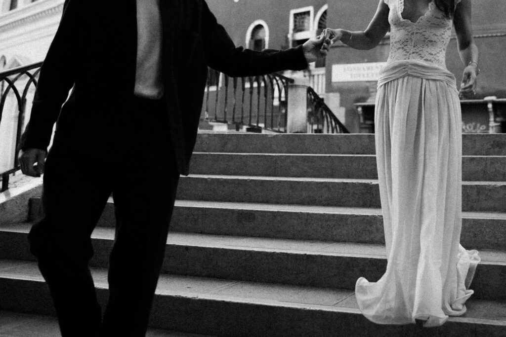 Black-and-white photograph of a couple holding hands as they descend stone bridge steps in Venice, showing her flowing ivory dress and his dark outfit.
