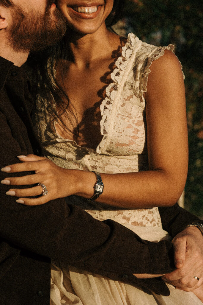 Close-up of a couple embracing in warm sunlight, showing her smile, ivory lace dress and rings against his dark brown outfit.
