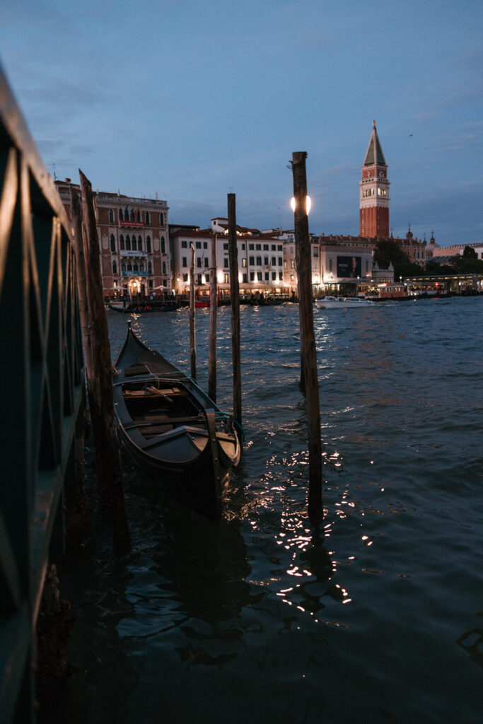 Gondola moored beside wooden posts at dusk in Venice, with waterfront lights reflected on the water and St Mark’s Campanile in the distance.
