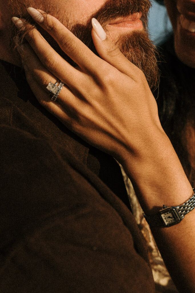 Close-up of a woman’s hand resting on her partner’s bearded cheek, with her rings and rectangular wristwatch in golden sunlight.