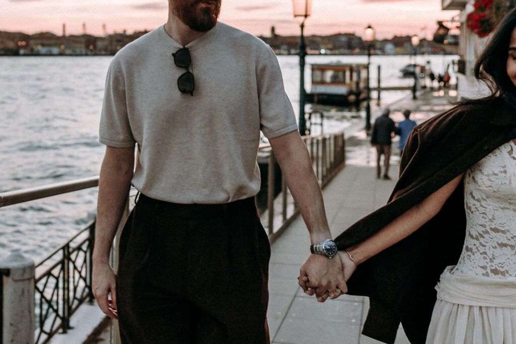 Newlyweds holding hands on a Venice waterfront at dusk, with his brown shirt draped over her shoulders and an older couple walking together in the background.
