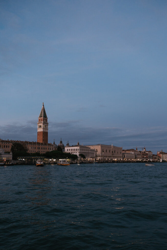 View across the Venetian lagoon towards St Mark’s Campanile and the Doge’s Palace at dusk, beneath a soft blue sky.