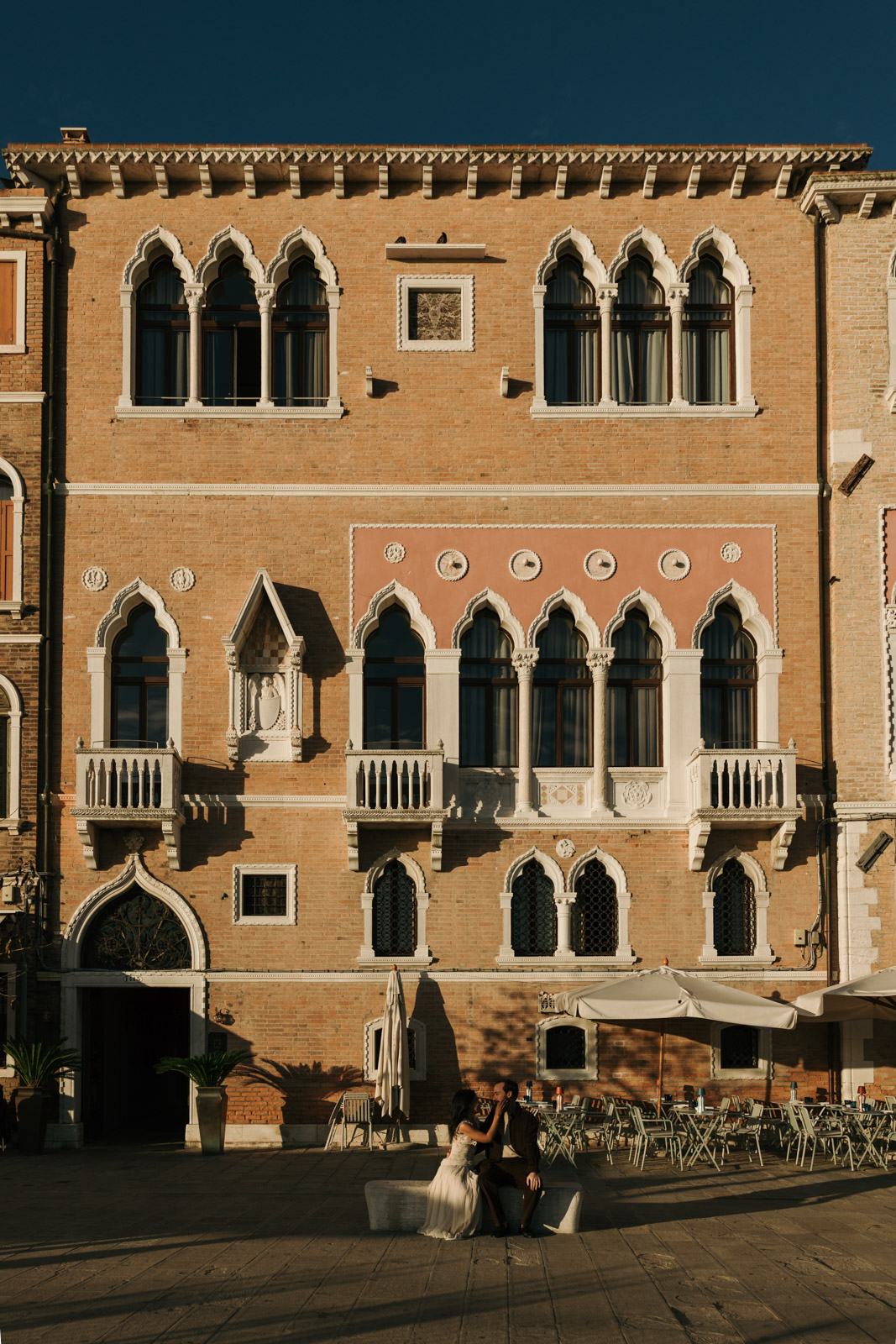 Couple sitting closely on a stone bench beneath an ornate brick façade with Gothic windows in Dorsoduro, Venice, in warm afternoon sunlight.