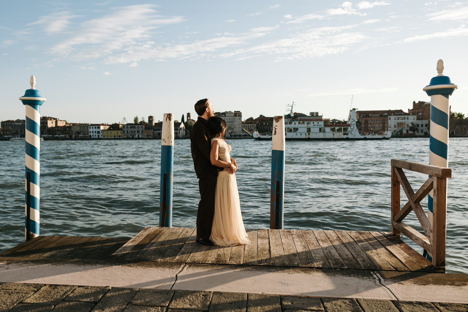 Couple embracing on a wooden dock in Dorsoduro, Venice, framed by blue-and-white mooring poles in afternoon sunlight.